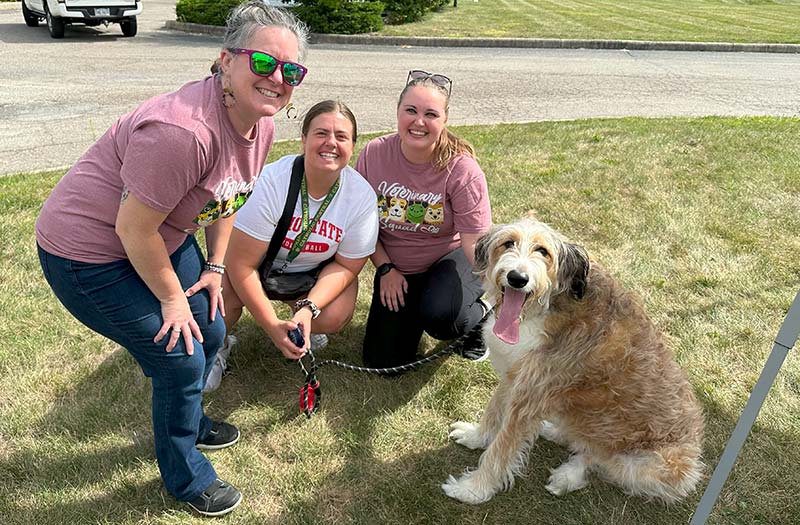 Three Happy Paws & Claws staff crouched on the lawn beside a shaggy, happy dog in Englewood, OH